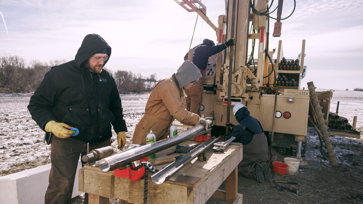 Workers operate drilling equipment in a snowy field, focusing on machinery and tools.