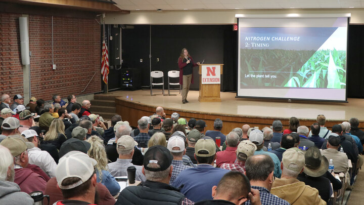 Speaker presenting to a seated audience in a lecture hall, slideshow on agriculture displayed.